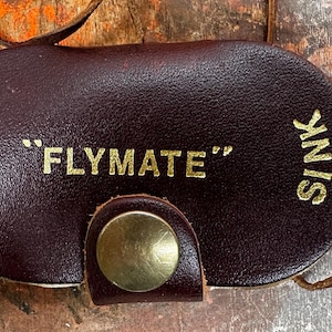 May include: A dark brown leather oval case with a gold-coloured metal snap closure and a thin leather cord. The text "FLYMATE" is printed in gold in the centre, with the word "FLOAT" on the left and "SINK" on the right side of the case.