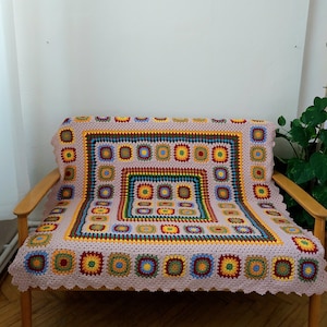 Puede incluir: Una manta de ganchillo colorida con un patrón geométrico en tonos de rosa, amarillo, verde, azul y rojo. La manta está colocada sobre un sofá de madera.