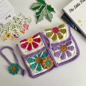 Könnte beinhalten: Gehäkelte Clutch mit Blumenmuster. Die Tasche hat einen weißen Grund mit lila Rand und bunte Blumen in Rosa, Gelb, Grün und Türkis. Ein passender Schlüsselanhänger mit Blume ist ebenfalls enthalten.