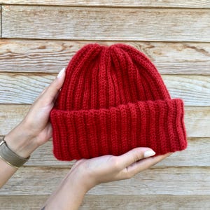 May include: A red knitted beanie hat with a ribbed texture. The hat is held in two hands.