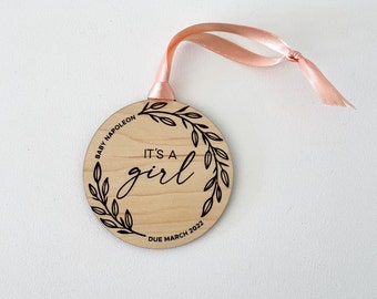 May include: A wooden round ornament with a pink ribbon. It has a floral design and the text "It's a girl" with the name "Baby Napoleon" and the date "Due March 2022".