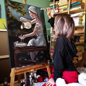 May include: A woman in a white dress and a white cap is painting a canvas with a scene of a woman pouring water from a pitcher. The woman in the painting is holding a small bird in her hand. The artist is sitting on the floor with a white cat beside her.