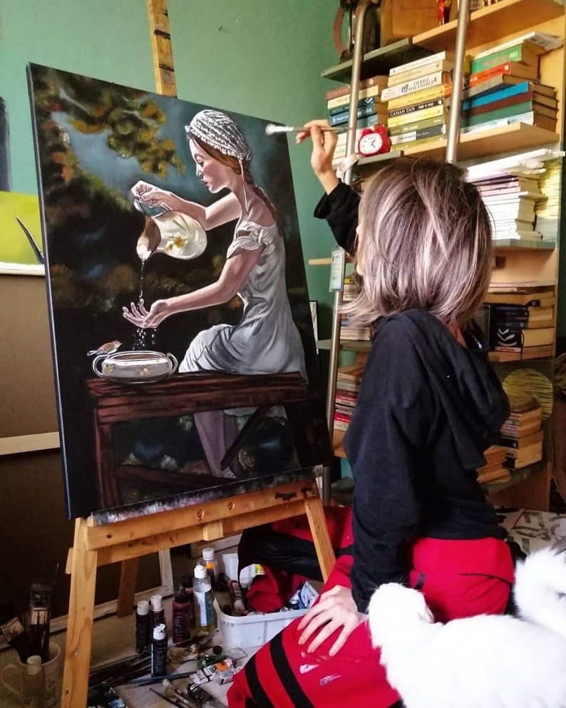 May include: A woman in a white dress and a white cap is painting a canvas with a scene of a woman pouring water from a pitcher. The woman in the painting is holding a small bird in her hand. The artist is sitting on the floor with a white cat beside her.