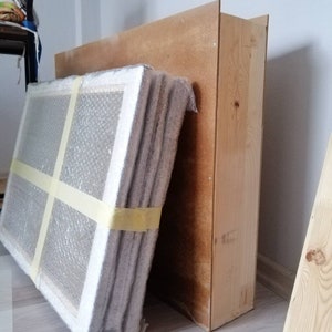 May include: A stack of wrapped canvases leaning against a wooden box. The canvases are wrapped in bubble wrap and secured with tape.