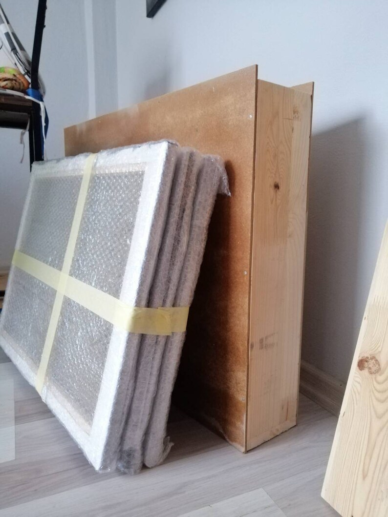 May include: A stack of wrapped canvases leaning against a wooden box. The canvases are wrapped in bubble wrap and secured with tape.