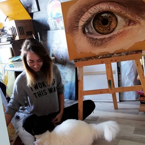Pu&ograve; includere: Una donna con una felpa grigia con la scritta "I woke up this way" &egrave; seduta sul pavimento con un gatto bianco. Dietro di lei, un cavalletto con un dipinto di un occhio grande.
