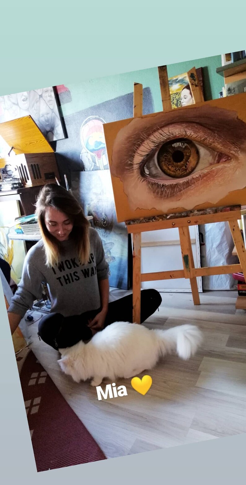 Pu&ograve; includere: Una donna con una felpa grigia con la scritta "I woke up this way" &egrave; seduta sul pavimento con un gatto bianco. Dietro di lei, un cavalletto con un dipinto di un occhio grande.
