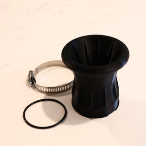 Op de afbeelding: Een zwart, trechtervormig onderdeel met een geribbeld ontwerp, vergezeld van een metalen klem en een zwarte O-ring. Het onderdeel heeft een brede opening en loopt taps toe naar de basis. De items zijn gerangschikt op een wit oppervlak.