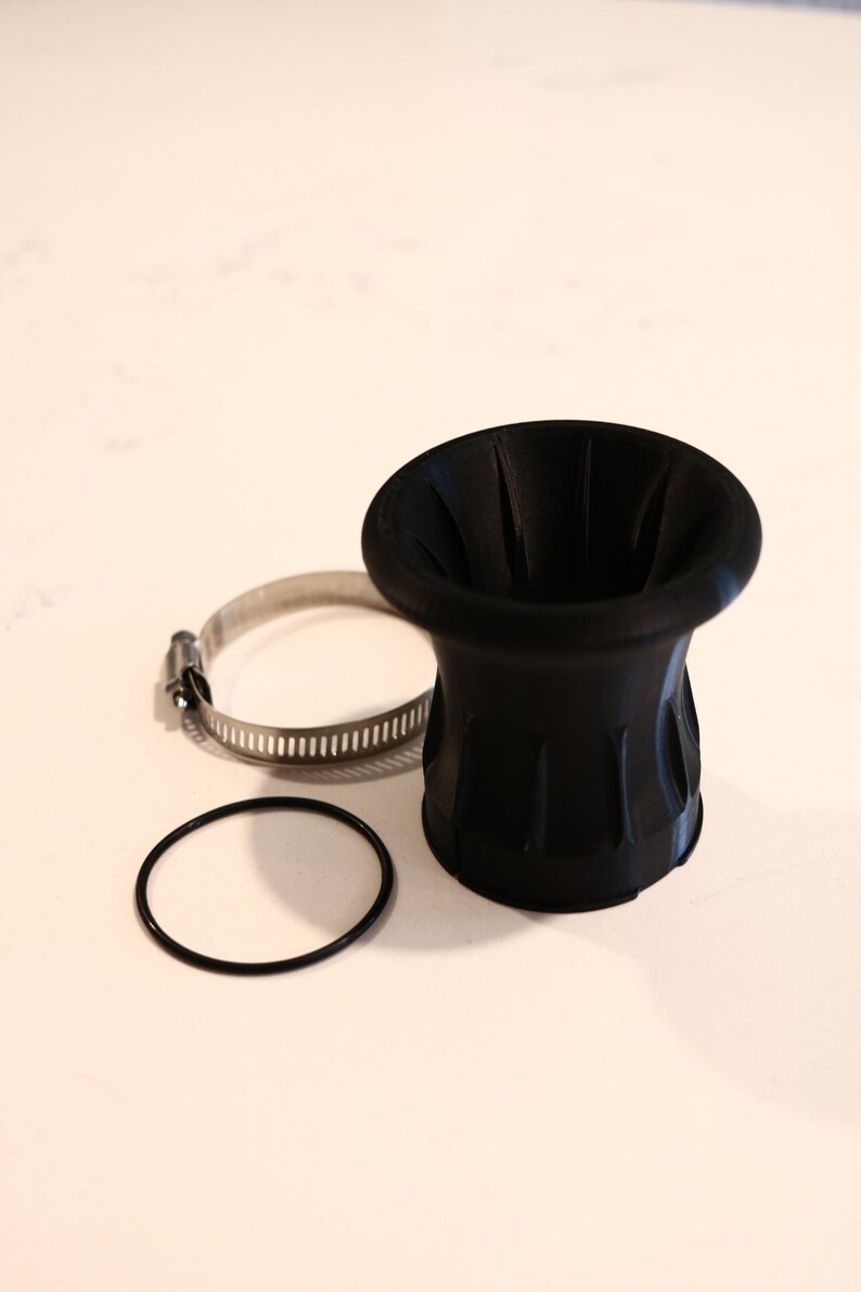 Op de afbeelding: Een zwart, trechtervormig onderdeel met een geribbeld ontwerp, vergezeld van een metalen klem en een zwarte O-ring. Het onderdeel heeft een brede opening en loopt taps toe naar de basis. De items zijn gerangschikt op een wit oppervlak.