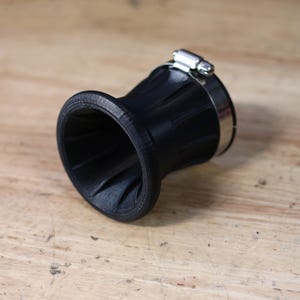 Op de afbeelding: Een zwart, trechtervormig object met een geribbelde buitenkant en een roestvrijstalen klem. De binnenkant van de trechter heeft een spiraalvormig ontwerp. Het object staat op een lichtbruin houten oppervlak.