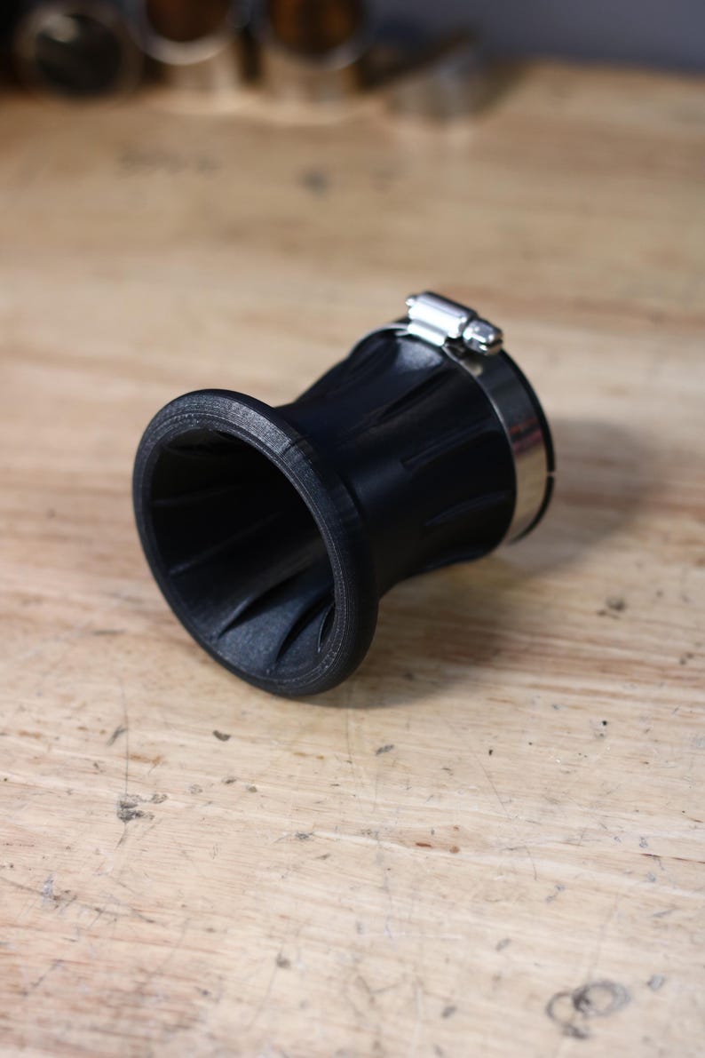 Op de afbeelding: Een zwart, trechtervormig object met een geribbelde buitenkant en een roestvrijstalen klem. De binnenkant van de trechter heeft een spiraalvormig ontwerp. Het object staat op een lichtbruin houten oppervlak.