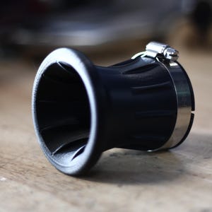 Op de afbeelding: Een zwart, trechtervormig object met een zilveren klem. Het object heeft een brede, uitlopende opening en een geribbelde buitenkant. De klem is gemaakt van glanzend metaal en is rond het lichaam bevestigd. Het ligt op een houten oppervlak.
