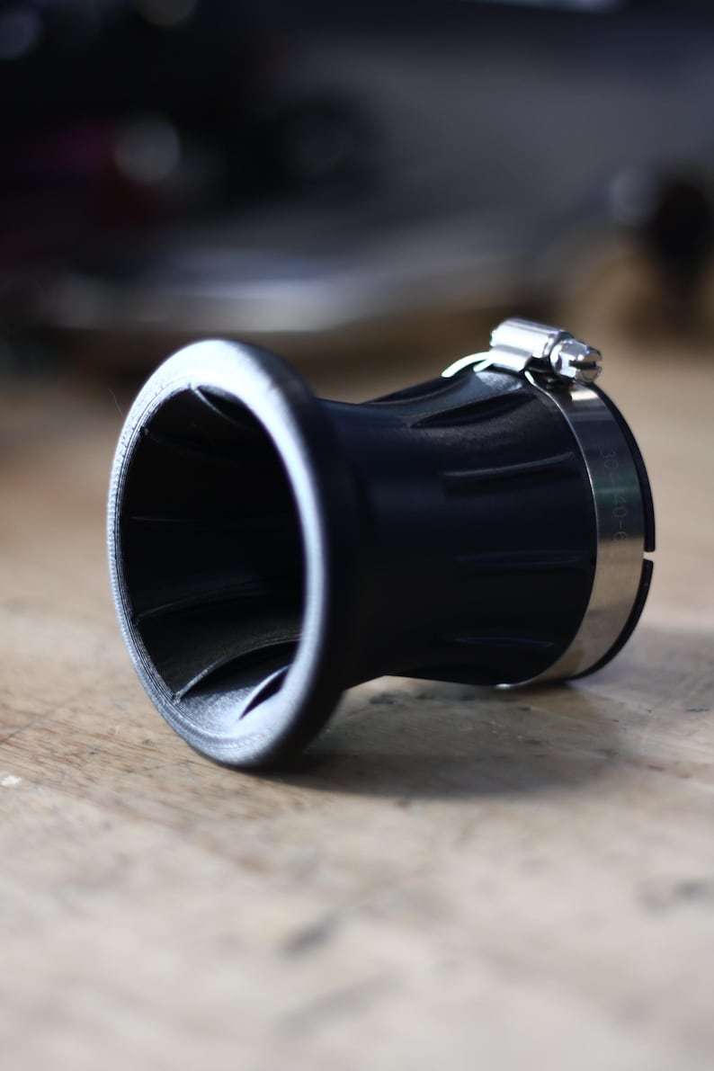 Op de afbeelding: Een zwart, trechtervormig object met een zilveren klem. Het object heeft een brede, uitlopende opening en een geribbelde buitenkant. De klem is gemaakt van glanzend metaal en is rond het lichaam bevestigd. Het ligt op een houten oppervlak.