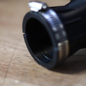 Op de afbeelding: Close-up van een zwarte rubberen slangconnector met een metalen klem. De connector heeft een cirkelvormige opening en een glad, gebogen ontwerp. De metalen klem is zilverkleurig en zet de slang vast. De achtergrond is een lichtbruin houten oppervlak.