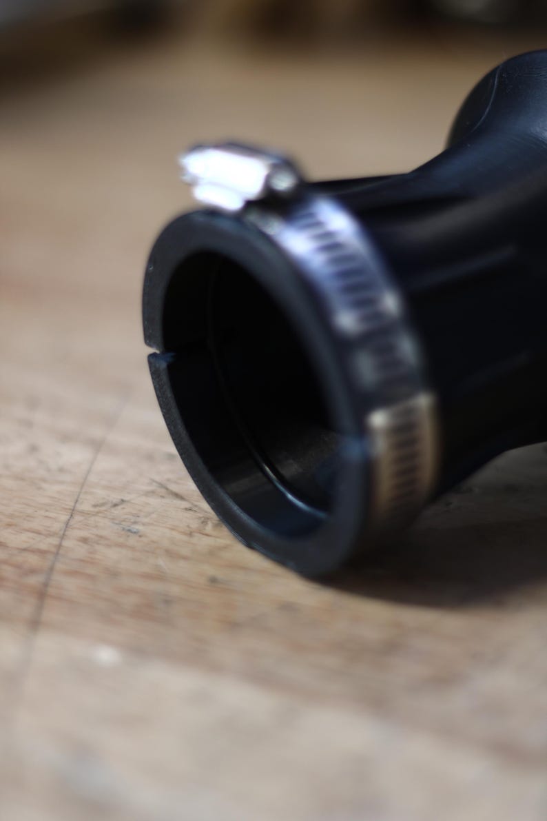 Op de afbeelding: Close-up van een zwarte rubberen slangconnector met een metalen klem. De connector heeft een cirkelvormige opening en een glad, gebogen ontwerp. De metalen klem is zilverkleurig en zet de slang vast. De achtergrond is een lichtbruin houten oppervlak.