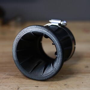 Op de afbeelding: Een zwart, trechtervormig object met een geribbelde binnenkant en een metalen klem. Het object lijkt van plastic te zijn en ligt op een houten oppervlak. De klem is zilverkleurig en glanzend.