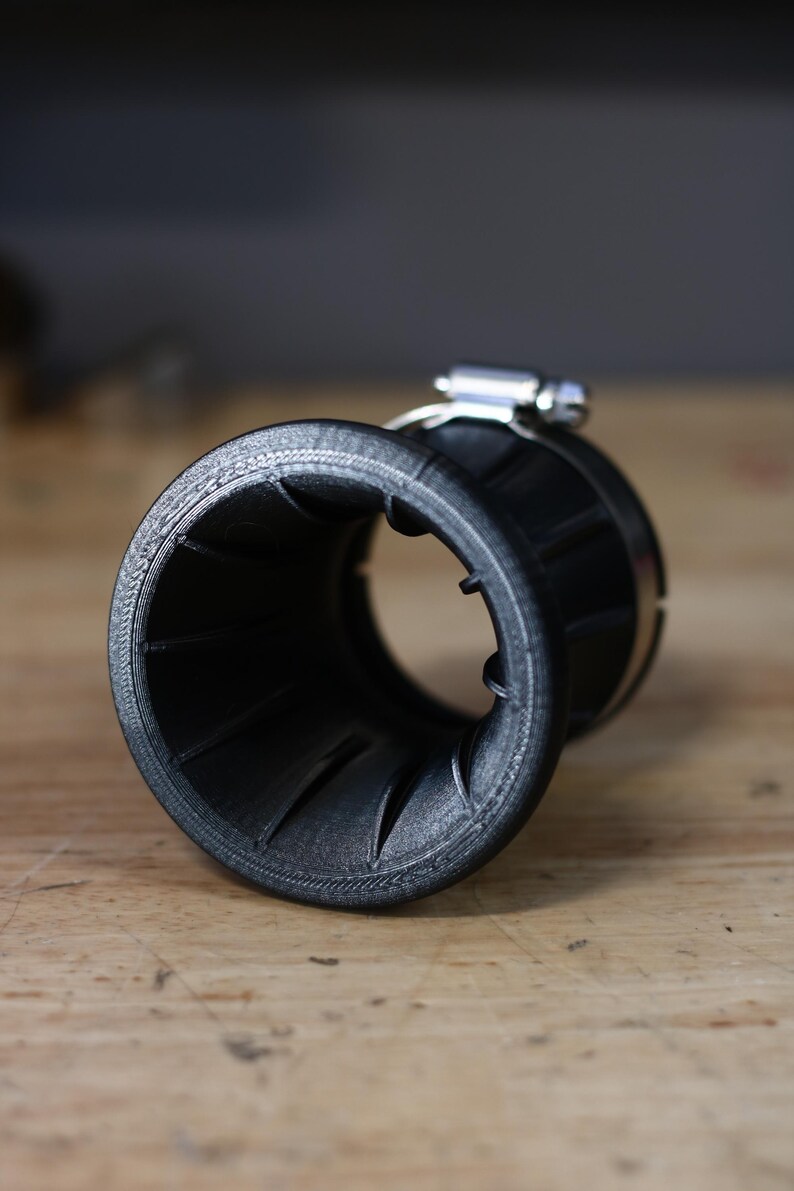 Op de afbeelding: Een zwart, trechtervormig object met een geribbelde binnenkant en een metalen klem. Het object lijkt van plastic te zijn en ligt op een houten oppervlak. De klem is zilverkleurig en glanzend.