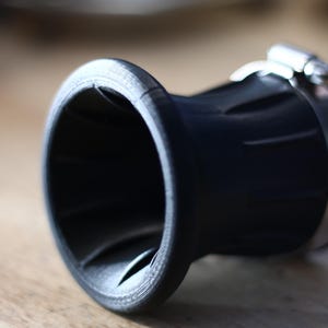 Op de afbeelding: Close-up van een zwart, cilindrisch object met een uitlopende opening. Het object heeft een getextureerd oppervlak en is vastgezet met een metalen klem. Het ontwerp suggereert een mechanisch of industrieel onderdeel.