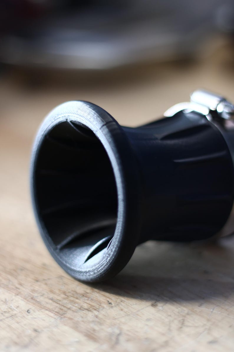 Op de afbeelding: Close-up van een zwart, cilindrisch object met een uitlopende opening. Het object heeft een getextureerd oppervlak en is vastgezet met een metalen klem. Het ontwerp suggereert een mechanisch of industrieel onderdeel.