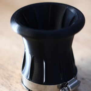 Op de afbeelding: Zwart trechtervormig object met een roestvrijstalen klem. Het object heeft een brede, uitlopende bovenkant en een geribbeld ontwerp aan de zijkanten. De klem is vastgezet met een schroef. Het object staat op een houten oppervlak.