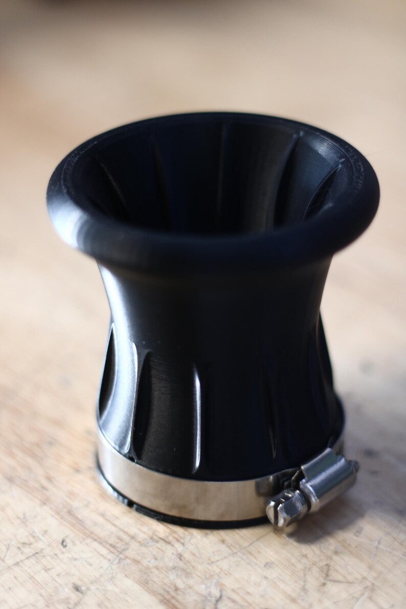 Op de afbeelding: Zwart trechtervormig object met een roestvrijstalen klem. Het object heeft een brede, uitlopende bovenkant en een geribbeld ontwerp aan de zijkanten. De klem is vastgezet met een schroef. Het object staat op een houten oppervlak.