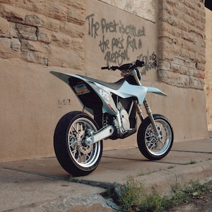 May include: A light blue supermoto motorcycle with a high-clearance frame, spoked wheels, and black tires. The bike features a rear license plate, a prominent number 93 graphic on the side panel, and a sleek, minimalist body design parked outdoors.