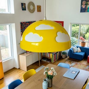 May include: A large, dome-shaped pendant light fitting with a glossy yellow finish and white cloud-shaped cutouts. The lamp hangs from a central rod above a wooden dining table, featuring a smooth, rounded surface and a wide, circular opening.