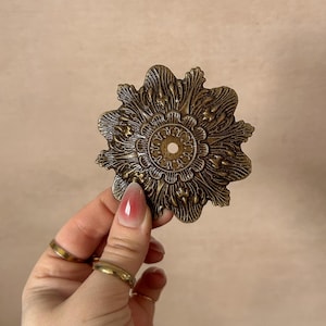 May include: Ornate, antique-style brass decorative rosette. The rosette features a detailed floral design with a central hole. The brass has a warm, aged patina. The rosette is held by a hand with gold rings.