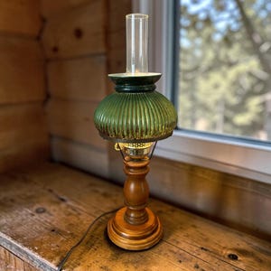 May include: A table lamp featuring a wooden base with a turned pedestal design, topped with a ribbed, emerald green glass shade and a clear glass chimney. The lamp includes a metallic burner assembly and a black electrical cord extending from the base.