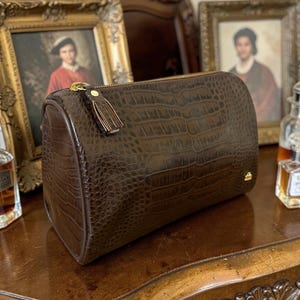 May include: A brown, barrel-shaped handbag with a crocodile skin texture. The bag has a gold zipper and a small tassel. The bag is sitting on a wooden surface. The background includes framed portraits and perfume bottles.