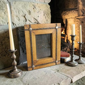 May include: A rectangular wooden picture frame with metal studs and a dark interior. Two brass candle holders with lit candles are on a stone surface. A stack of books is visible.