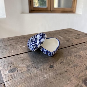May include: A heart-shaped ceramic trinket box with a lid, decorated with blue and white geometric patterns. The box is open, showing a white interior. It rests on a weathered wooden surface.
