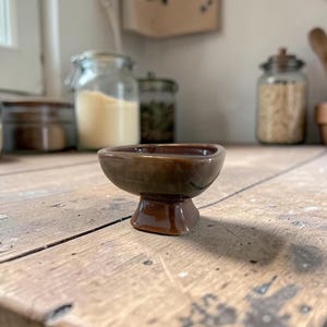 May include: A small, brown ceramic bowl with a pedestal base. The bowl has a rounded shape and a glossy finish. It sits on a rustic wooden surface, with jars and kitchen utensils in the background. The bowl is likely for serving small portions.