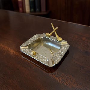May include: A rectangular silver-toned metal ashtray featuring an ornate embossed floral border. The ashtray is decorated with a small gold-toned golf ball and two crossed gold-toned golf clubs attached to the rim of the tray.