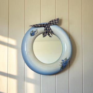 May include: A round wall mirror with a white and blue gradient frame featuring painted blue floral clusters on the sides. A blue and beige checkered fabric bow is attached to the top of the circular frame, adding a decorative accent to the piece.