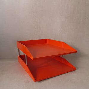 Peut inclure: Un organiseur de bureau orange à deux niveaux au design moderne. Les plateaux sont rectangulaires avec des côtés inclinés et sont soutenus par des poteaux métalliques argentés. L'organisateur est placé sur un fond neutre.