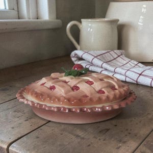 Puede incluir: Una fuente de cerámica para pastel con una parte superior de celosía y un borde decorativo. La fuente para pastel es de color rosa claro con detalles en rojo y una cereza verde y roja en la parte superior. El plato está sobre una mesa de madera.