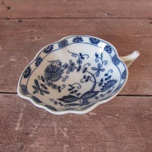 Puede incluir: Un plato blanco de porcelana con forma de hoja y un estampado floral azul. El plato tiene un borde festoneado y un pequeño asa. El diseño azul presenta flores, hojas y enredaderas. El plato es probablemente para servir o exhibir.