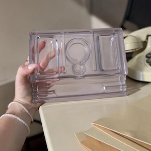 May include: A clear, rectangular plastic organiser with several compartments. It features a central circular section and two side compartments. The design is simple and functional, suitable for storing small items. The organiser is held by a person.