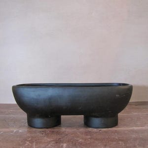 May include: A black, oblong planter with rounded sides and two cylindrical legs. The planter has a rectangular opening and sits on a wooden surface. The minimalist design is suitable for various plants.