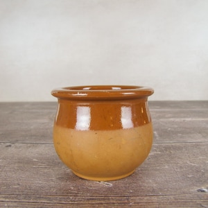 May include: A small, round, two-tone ceramic pot. The top section is a deep brown, fading into a lighter, tan base. The pot has a slightly flared rim and is placed on a wooden surface, giving a rustic or vintage feel.
