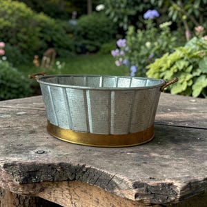 May include: An oval galvanised metal basin featuring vertical ribbed detailing and a smooth, solid brass-toned base. The container includes two small, curved metal handles attached to opposite sides, providing a functional grip for this vessel.