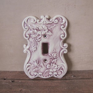 Könnte beinhalten: Eine dekorative Lichtschalterabdeckung in einem sanften Lavendelton. Die Keramikabdeckung hat ein Blumendesign mit erhabenen Reliefdetails. Die Kanten sind mit Schnörkeln verziert. Die Schalteröffnung hat eine Standardgröße.