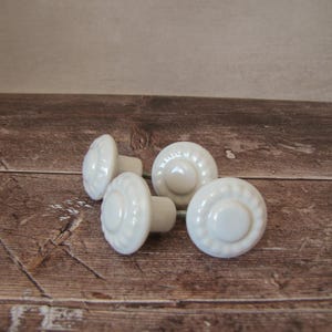 Peut inclure: Quatre boutons de meuble en céramique blanche avec un motif floral. Les boutons sont ronds avec un centre légèrement bombé et un bord décoratif. Ils sont disposés sur une surface en bois.