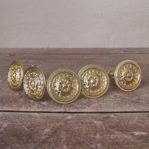 Pode incluir: Seis puxadores de porta antigos de latão com um design floral. Os puxadores estão dispostos sobre uma superfície de madeira. Os puxadores são de cor dourada brilhante e têm um padrão detalhado em relevo.