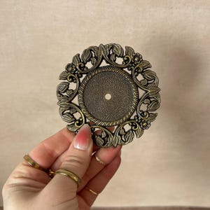 May include: An antique bronze metal decorative piece with an ornate design. The circular piece has a textured central area with a small hole, framed by a detailed floral and leaf pattern. It has a vintage aesthetic.