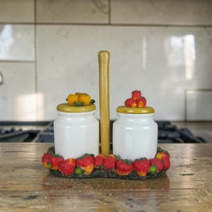 May include: A white ceramic salt and pepper shaker set with a wooden handle. The shakers have yellow lids and are decorated with a ring of red strawberries. The set is on a wooden surface.