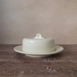 Pode incluir: Uma manteigueira de cerâmica branca com tampa e uma pequena pega. O prato está sobre um prato a condizer. A manteigueira está sobre uma superfície de madeira.