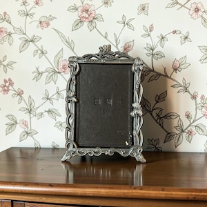 Peut inclure: Un cadre photo rectangulaire avec un fond marron foncé et un bord argenté métallique orné de motifs floraux et de feuilles en relief. Le cadre repose sur quatre petits pieds et présente une crête décorative avec des détails botaniques.