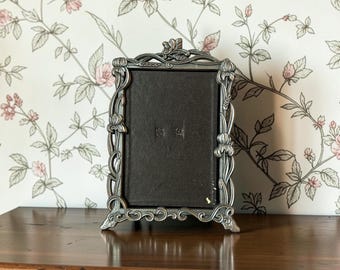 May include: A rectangular picture frame with a dark brown backing and a metallic silver-toned border featuring embossed floral and leaf motifs. The frame stands on four small feet and has a decorative top crest with sculpted botanical details.